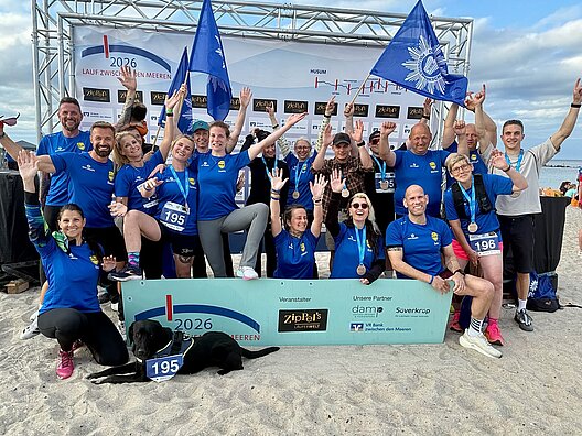 Gruppenfoto der DPolG Berlin nach dem erfolgreichen „Lauf zwischen den Meeren 2026“ am Ostseestrand in Damp. Die Läuferinnen und Läufer tragen ihre Finisher-Medaillen, halten DPolG-Fahnen in die Höhe und jubeln gemeinsam vor der offiziellen Fotowand des Veranstalters. Im Vordergrund liegt die schwarze Labradorhündin Bella mit einer symbolischen Startnummer im Sand. Das Bild steht für Teamgeist, Zusammenhalt und die erfolgreiche Bewältigung der 92,2 Kilometer langen Staffel von Husum an der Nordsee bis nach Damp an der Ostsee.