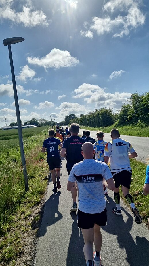 Teilnehmerinnen und Teilnehmer des „Lauf zwischen den Meeren 2026“ laufen auf einer asphaltierten Landstraße durch die schleswig-holsteinische Landschaft. Die Aufnahme zeigt das Läuferfeld von hinten, umgeben von grünen Feldern, Bäumen und einem sonnigen Himmel mit lockeren Wolken. Das Bild vermittelt die Weite der Strecke und die besondere Atmosphäre des 92,2 Kilometer langen Staffellaufs von Husum nach Damp.