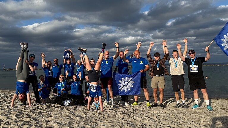 Gruppenfoto der DPolG Berlin nach dem „Lauf zwischen den Meeren 2026“ am Ostseestrand in Damp. Die Läuferinnen und Läufer stehen mit ihren Finisher-Medaillen im Sand, halten DPolG-Fahnen in die Höhe und jubeln gemeinsam. Zwei Teilnehmende machen einen Handstand, während die Gruppe fröhlich für das Erinnerungsfoto posiert. Im Hintergrund sind die Ostsee, ein bewölkter Himmel und die Küstenlandschaft zu sehen. Das Bild vermittelt Teamgeist, Freude und den erfolgreichen Abschluss der 92,2 Kilometer langen Staffel von Husum nach Damp.