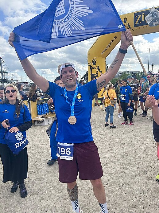 Ein Läufer der DPolG Berlin jubelt nach dem Zieleinlauf des „Lauf zwischen den Meeren 2026“ am Ostseestrand in Damp und hält eine große DPolG-Fahne über dem Kopf. Er trägt die Finisher-Medaille und die Startnummer 196. Im Hintergrund sind weitere Teilnehmerinnen und Teilnehmer sowie der gelbe Zielbogen des Veranstalters Zippel’s Läuferwelt zu sehen. Das Bild vermittelt Stolz, Freude und den erfolgreichen Abschluss der 92,2 Kilometer langen Staffel von Husum nach Damp.