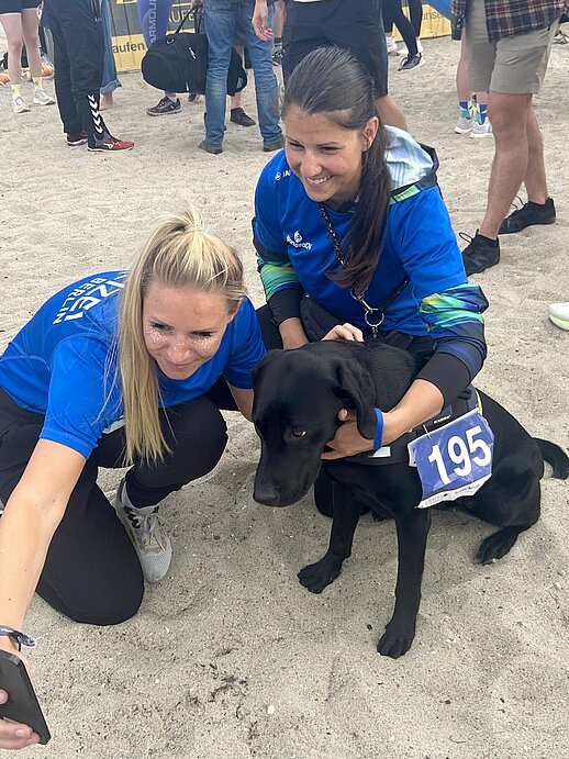 Zwei Läuferinnen der DPolG Berlin knien nach dem „Lauf zwischen den Meeren 2026“ am Ostseestrand in Damp neben der schwarzen Labradorhündin Bella. Die Hündin trägt symbolisch eine Startnummer, während die beiden Frauen in blauen Teamshirts lächelnd für ein Erinnerungsfoto posieren. Im Hintergrund sind weitere Teilnehmerinnen und Teilnehmer des Staffellaufs im Zielbereich zu sehen. Das Bild vermittelt Freude, Gemeinschaft und die entspannte Atmosphäre nach dem erfolgreichen Zieleinlauf.